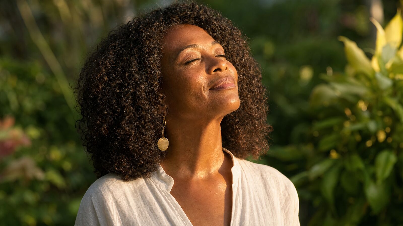 Woman with eyes closed, face tilted to warm sunlight in a green garden
